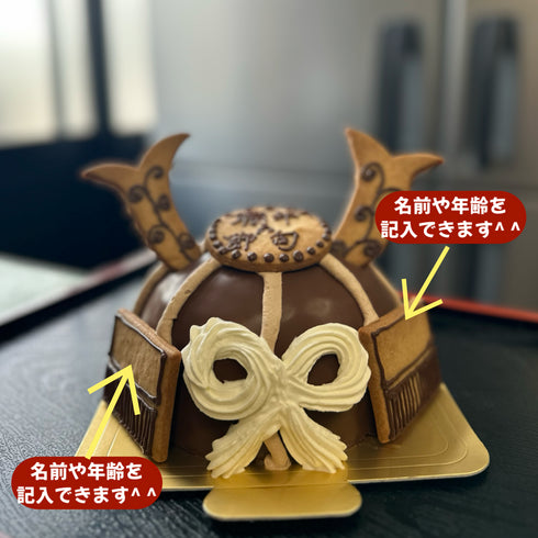 生チョコでコーティングし、クッキーや生クリームでデコレーションした兜ケーキの写真。左右のクッキーに名入れなど文字入れができることを説明している。